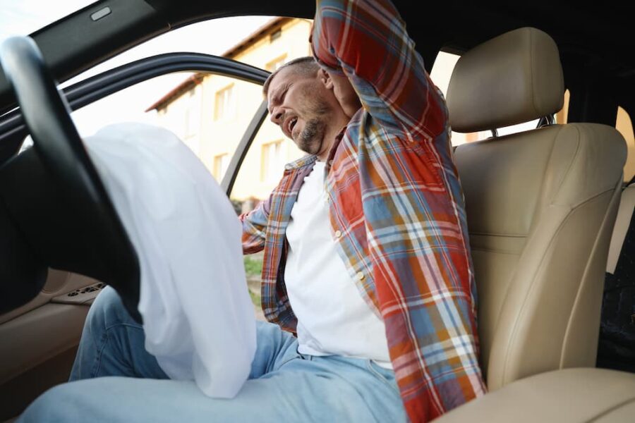 A man clutching his neck in pain inside a car with a deployed airbag, illustrating immediate steps for what to do after a car accident and seeking medical care.
